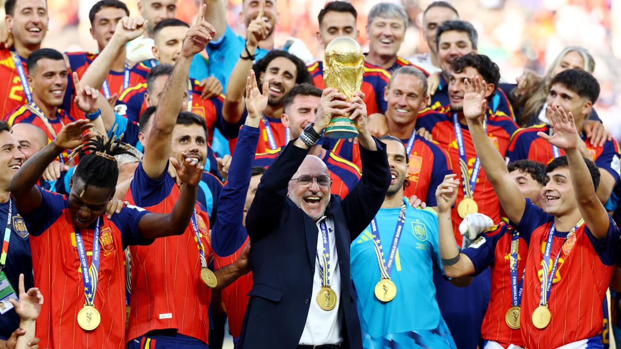 Spain national football team is celebrating its victory at the 2026 FIFA World Cup. Head coach Luis de la Fuente is prominently featured in the center, triumphantly lifting the World Cup trophy surrounded by his ecstatic players after defeating Argentina 1–0 in extra time. Image used for Sportxparte news.