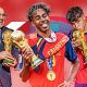A celebratory sports graphic featuring Spain's national football team after winning the 2026 FIFA World Cup, with manager Luis de la Fuente, forward Lamine Yamal sticking out his tongue, and defender Pau Cubarsí kissing the FIFA World Cup trophy, all wearing gold champion medals over their red and blue national jerseys against a vibrant red background. Image used for Sportxparte news.