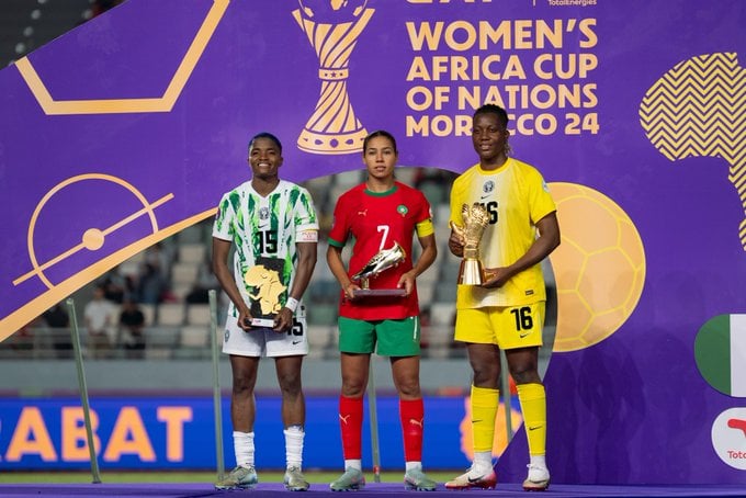 From left to right, Nigeria's Rasheedat Ajibade in a green-and-white zig-zag jersey holds a map-shaped MVP award; Morocco's Ghizlane Chebbak in a red shirt with green shorts holding a golden boot top scorer award; and Nigeria's goalkeeper Chiamaka Nnadozie in a bright yellow jersey holding a golden glove trophy at the 2024 Women's Africa Cup of Nations podium in Morocco. Image used for Sportxparte news.