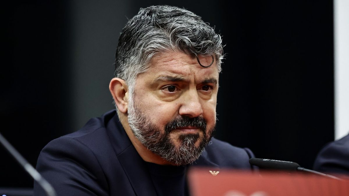 Italian football manager Gennaro Gattuso during a press conference. He is wearing a dark navy blue suit jacket over a dark T-shirt. He has salt-and-pepper hair and a thick, well-groomed beard, looking forward with a serious, emotional expression. Microphones are visible in the blurred foreground. Image used for Sportxparte news.