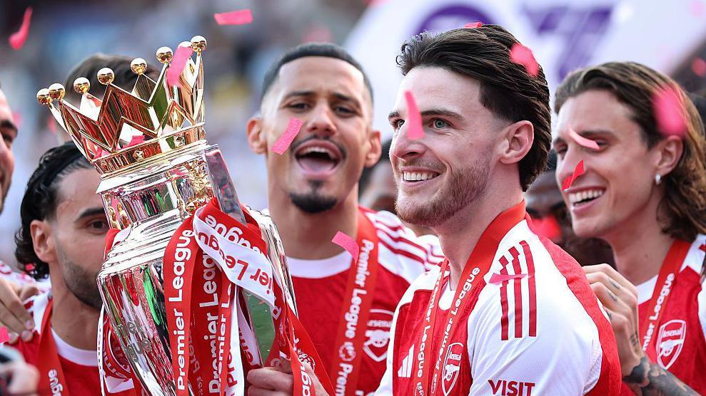 Arsenal midfielder Declan Rice celebrating with teammates after winning the Premier League trophy in May 2026, ending the club's 22-year wait for the league title. Image used for Sportxparte news.