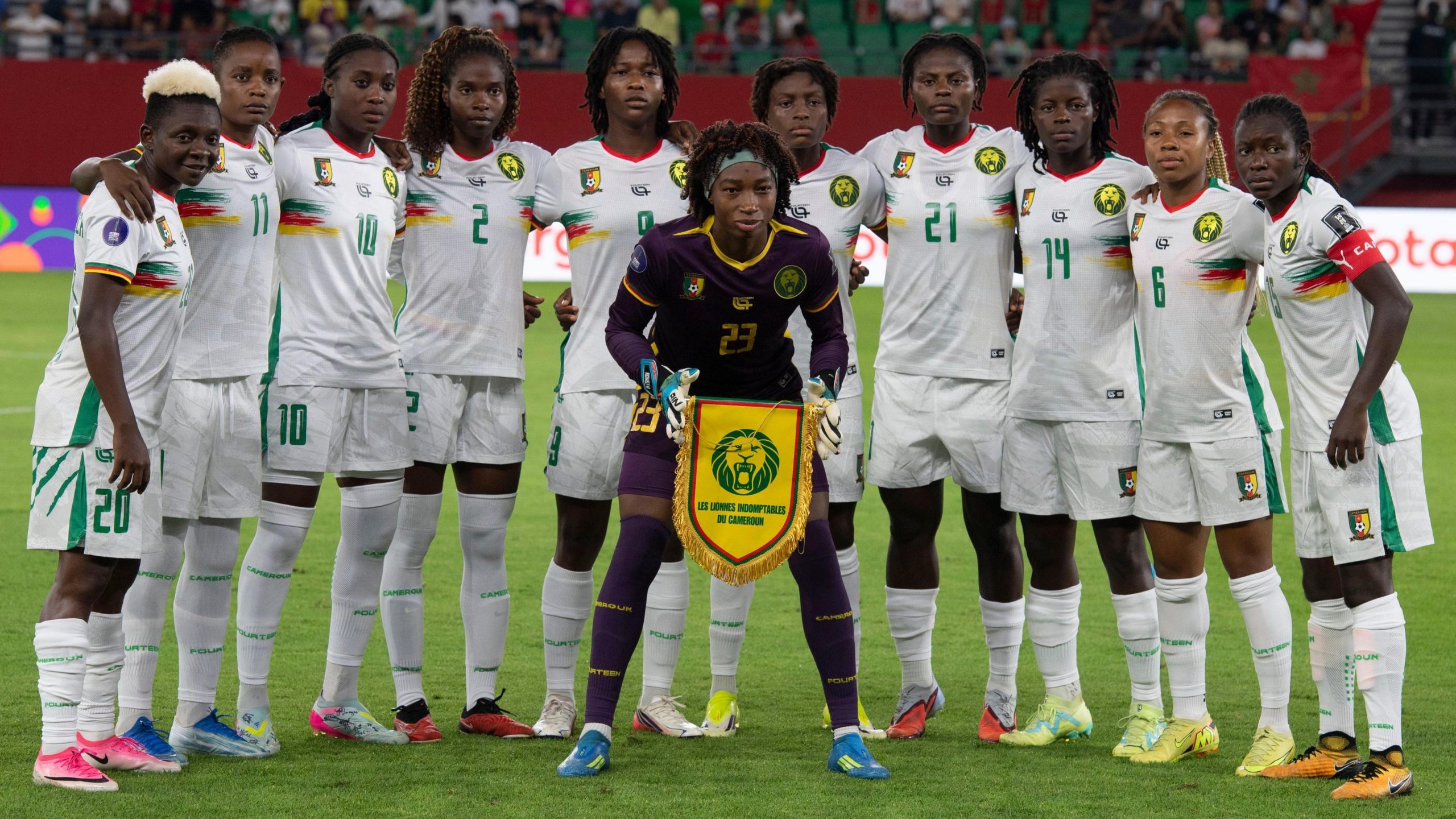 Cameroon women's national football team lineup standing on the pitch holding the Indomitable Lionesses team pennant banner. Image used for Sportxparte news.