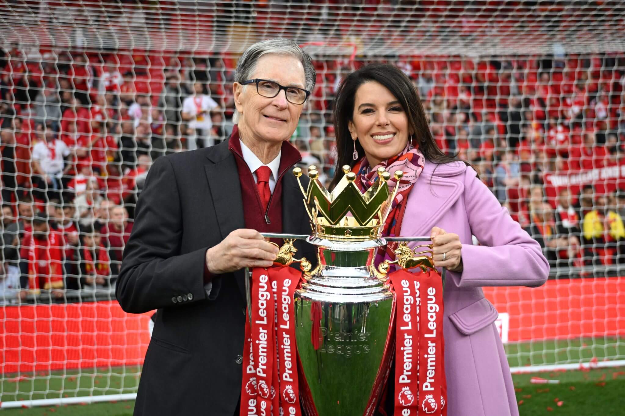 John W Henry and his wife Linda Pizzuti Henry, smiling while holding the English Premier League trophy together at Anfield Stadium. Image used for Sportxparte news.